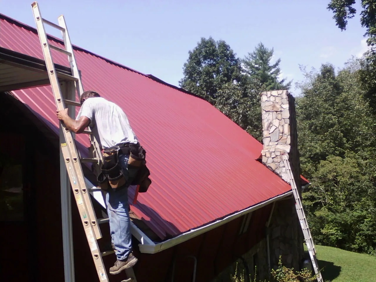 Roofer on a ladder beside a red metal roof and stone chimney — Soffit & Fascia Repair by Roofing Brothers in Shingletown
